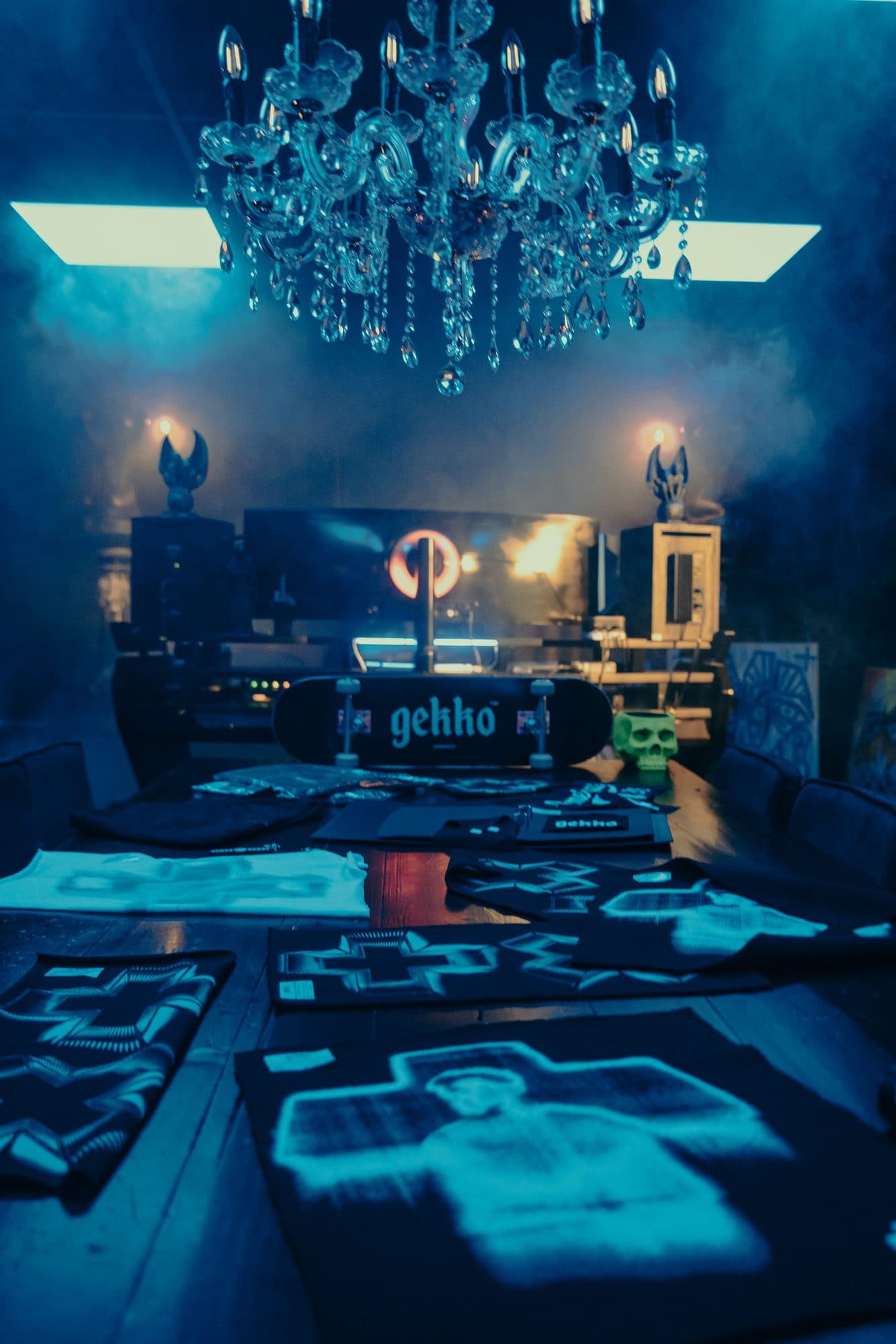 Symmetrical view down the creative table under a crystal chandelier, with a GEKKO skateboard, a green skull and printed sample panels in blue haze.