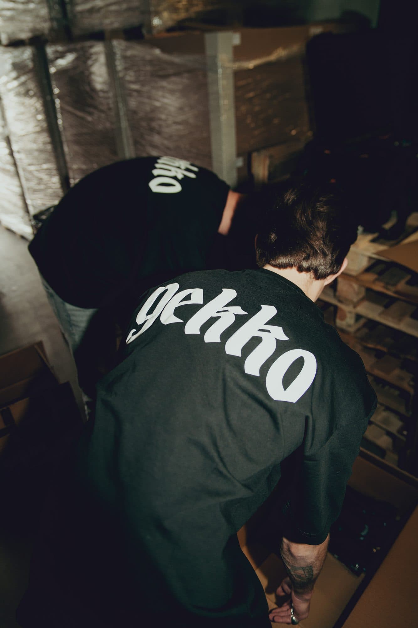 Two team members in black GEKKO tees seen from behind, bending over boxes between wrapped pallets in the warehouse.