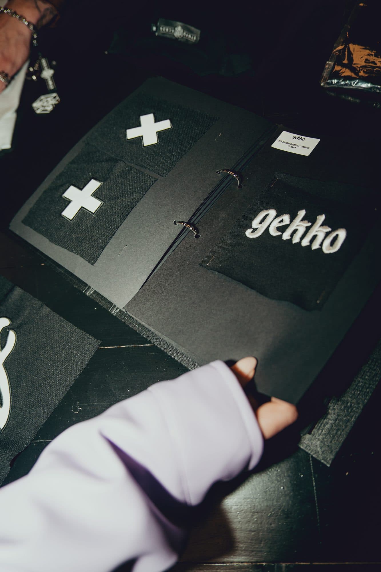 Hand in a lilac sleeve turning a page of an embroidery swatch book with white cross patches and a raised foam GEKKO logo.