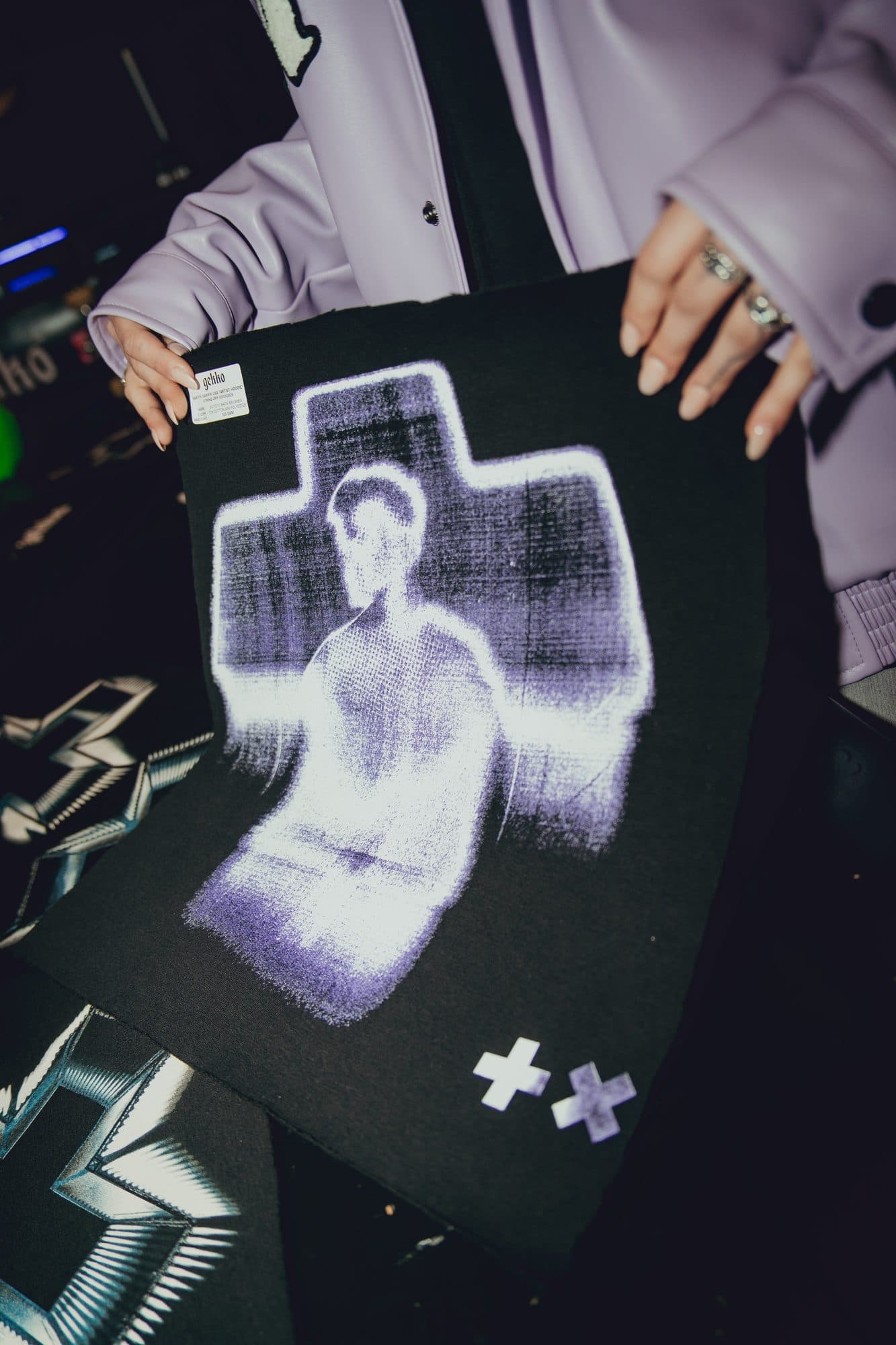 Hands in a lilac jacket holding up a black hoodie panel with a purple photographic print of a figure inside a cross.