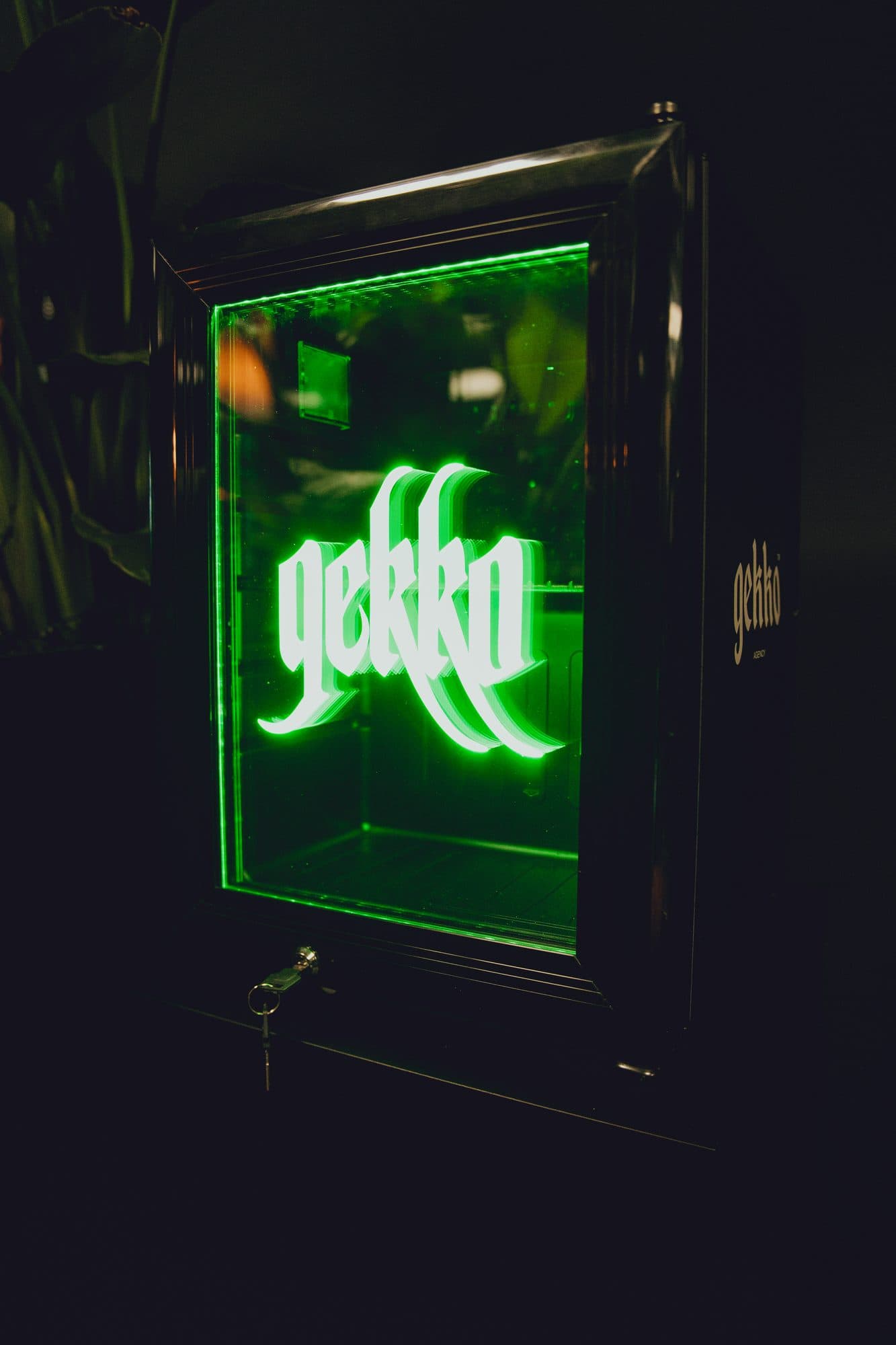 Black mini fridge with a glowing green neon GEKKO logo inside its glass door, a key in the lock and plant leaves at the left.