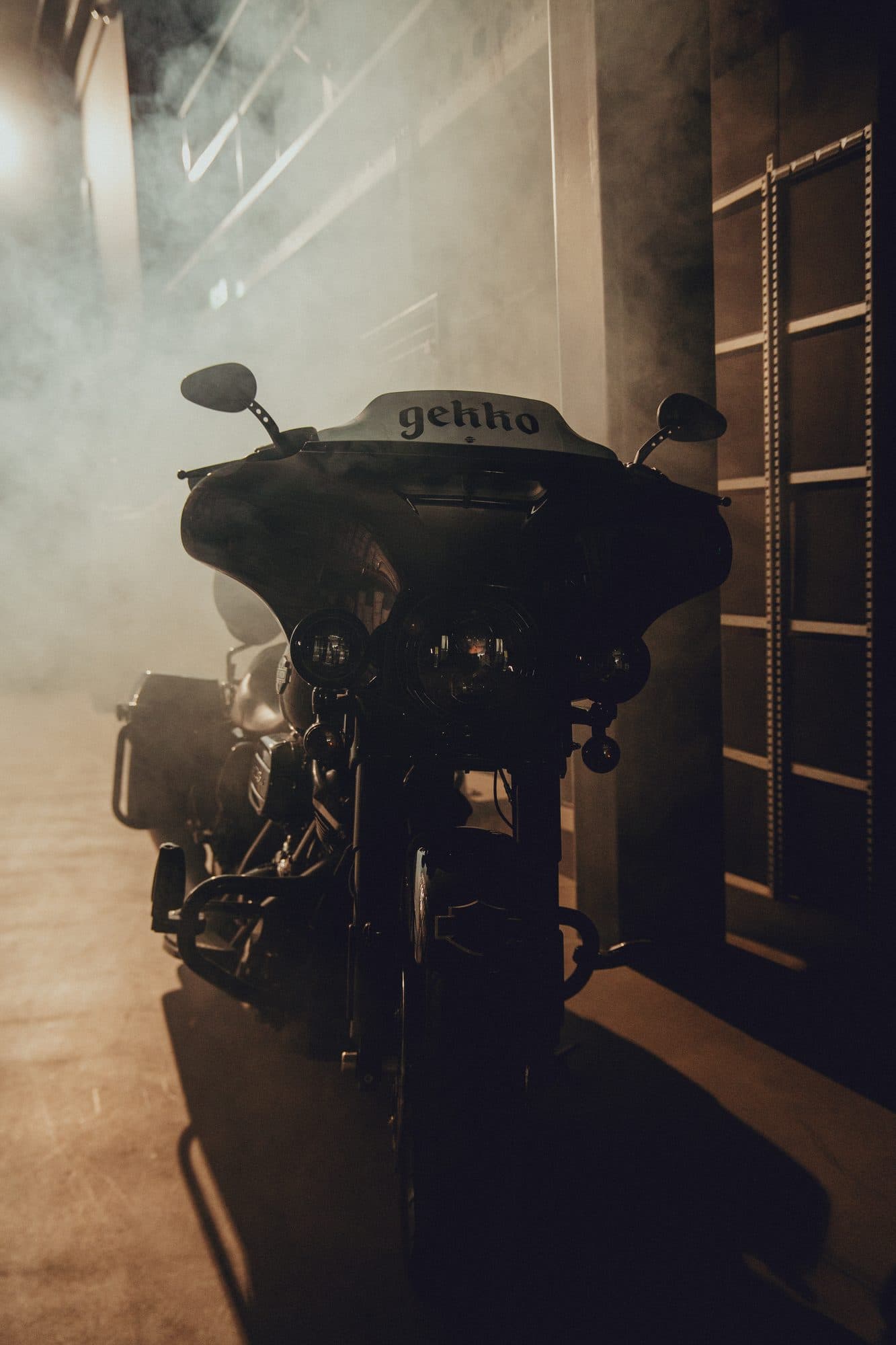 Black touring motorcycle with GEKKO on the windscreen standing in thick smoke between warehouse racks.