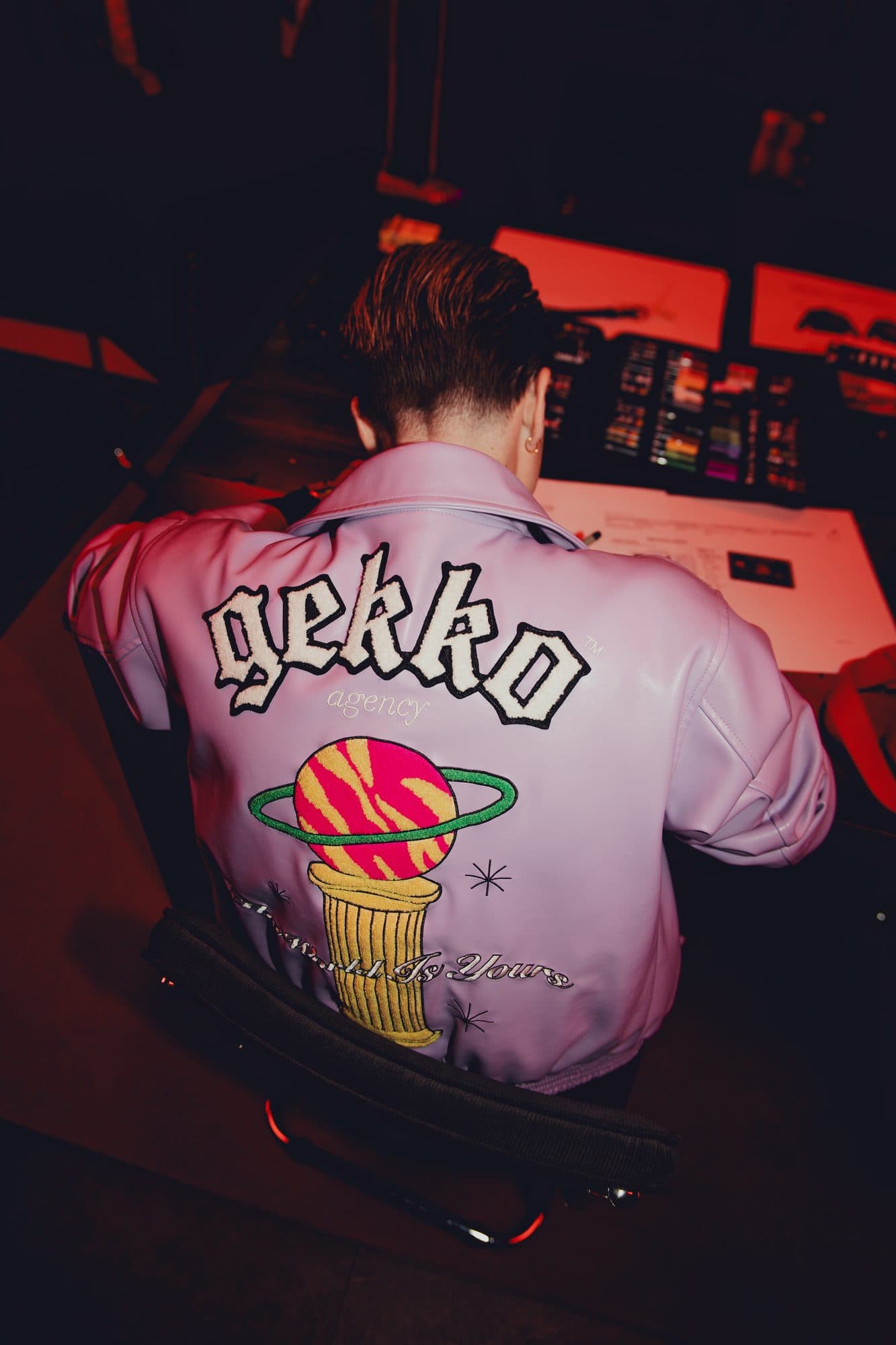 Designer seen from behind at a desk in red light, wearing a lilac leather jacket with a chenille GEKKO logo and an embroidered planet on a pedestal.