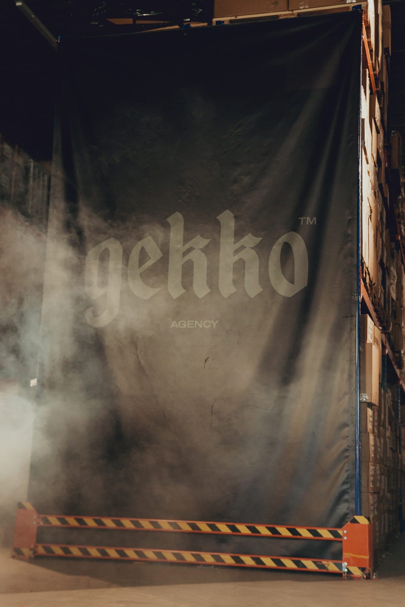 Large dark GEKKO Agency banner hanging over pallet racking in the warehouse, with smoke drifting past and a yellow and black striped safety barrier at the bottom.