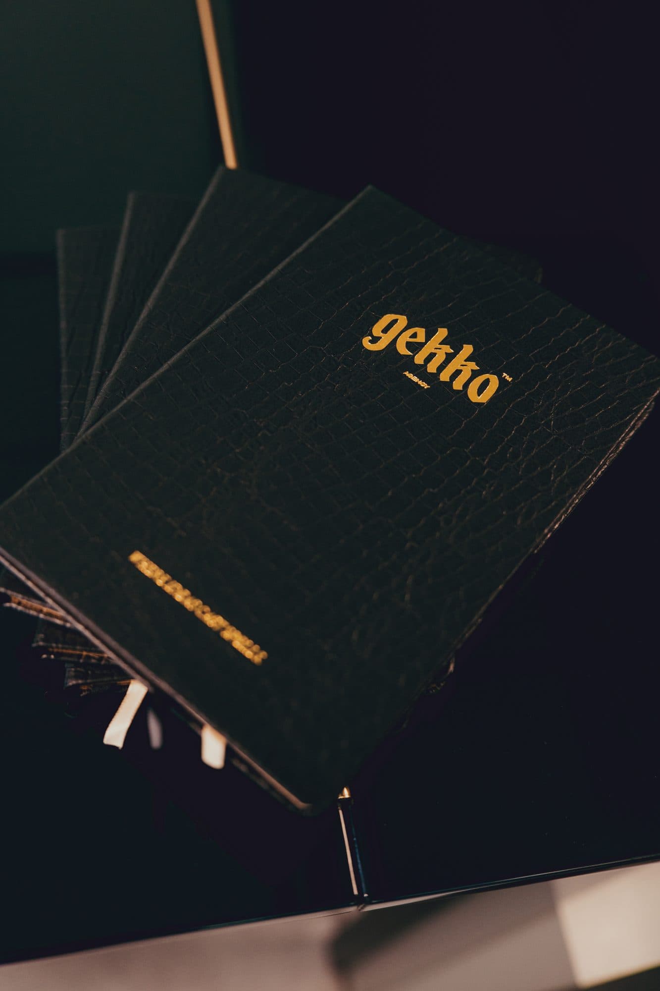 Fanned stack of black crocodile-embossed GEKKO notebooks with a gold foil logo and ribbon markers.