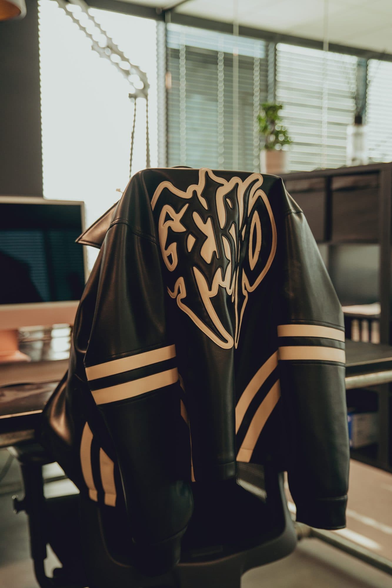 Black leather jacket with a cream GEKKO logo appliqué and striped sleeves draped over an office chair, with daylight through the blinds behind.