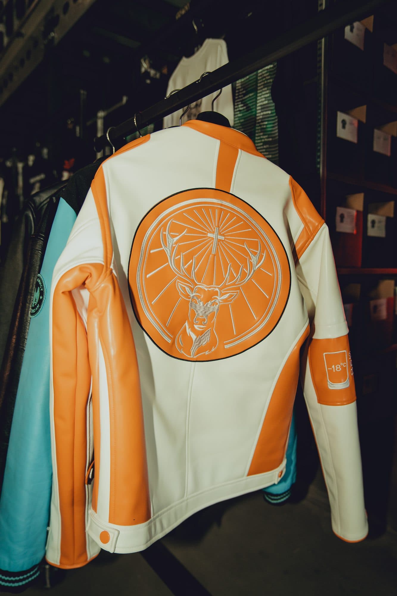 White and orange leather racing jacket with a large embroidered stag crest on the back, hanging on a rail in the warehouse.