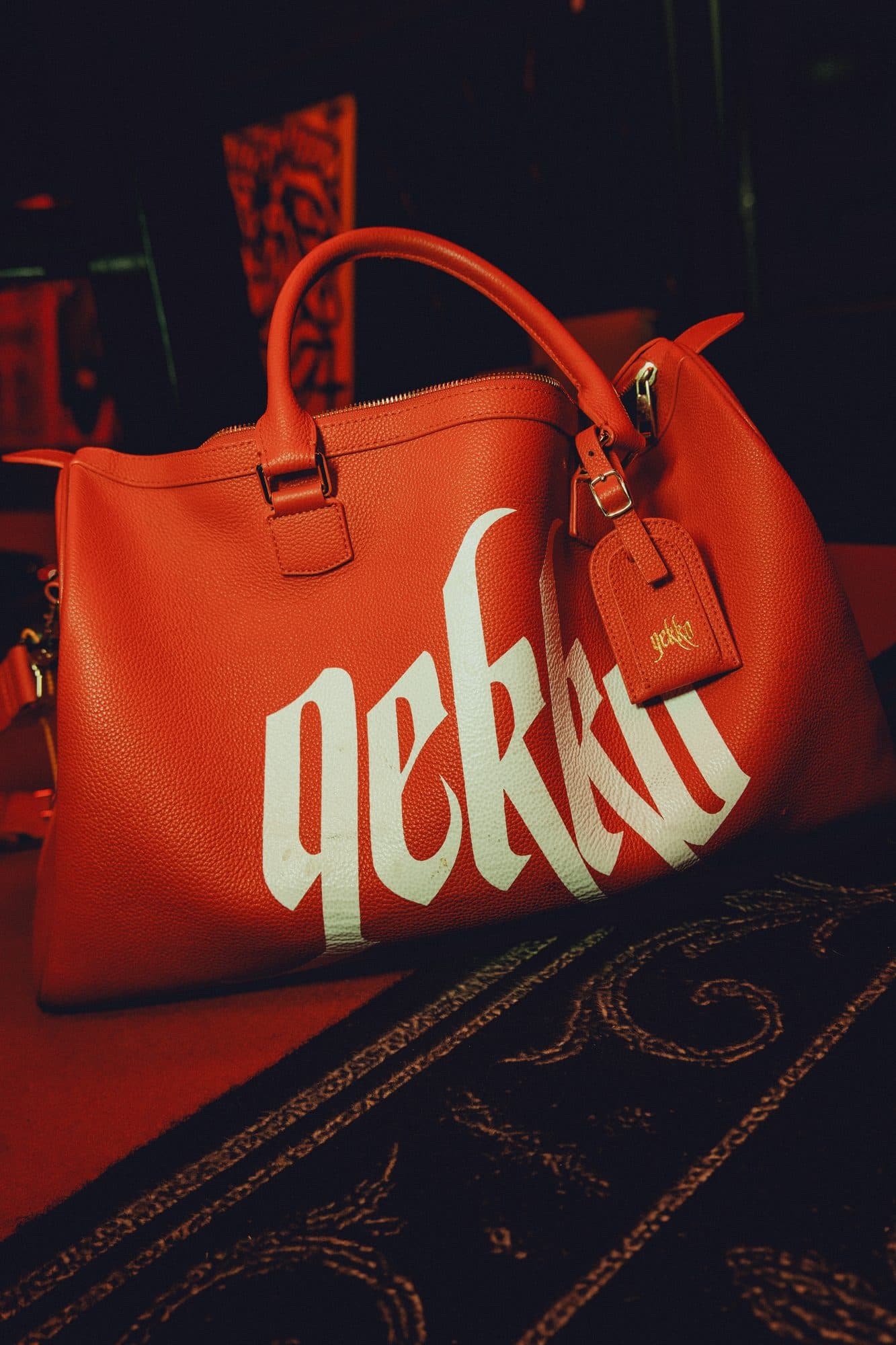 Red leather weekender bag with a large white GEKKO logo and a matching luggage tag on a dark patterned carpet.