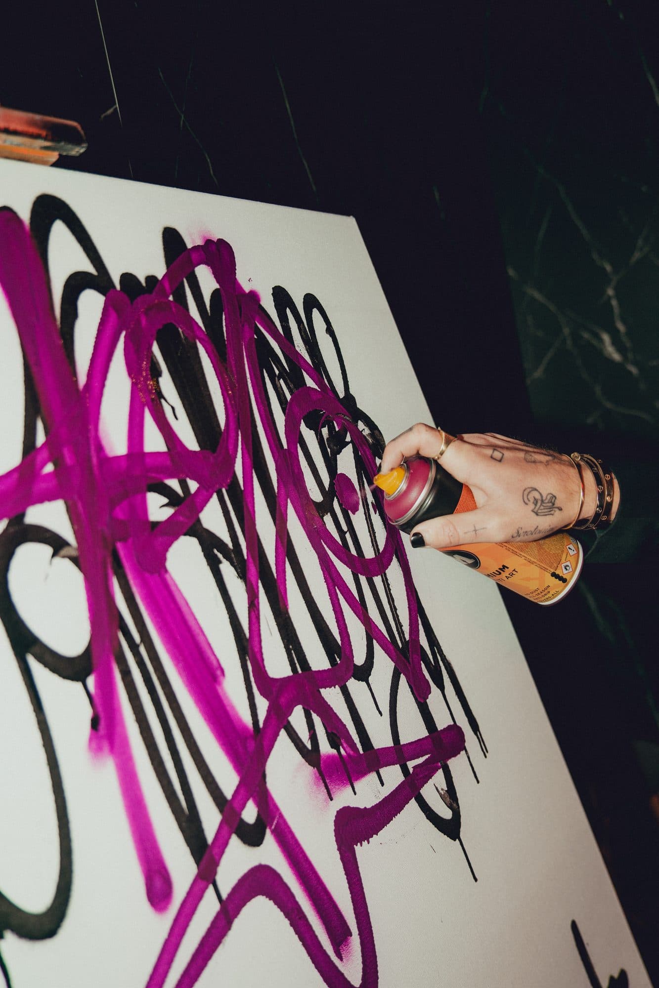 Tattooed hand with rings spraying magenta paint over black tags on a white canvas.
