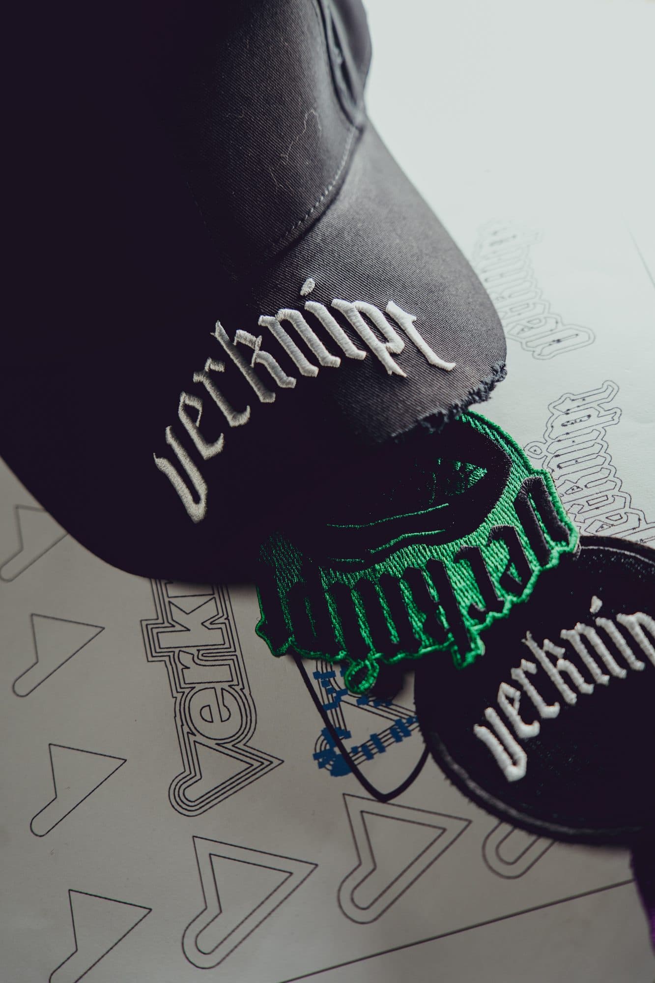 Raised white Verknipt embroidery on the brim of a distressed black cap, resting on embroidered Verknipt patches and artwork printouts.