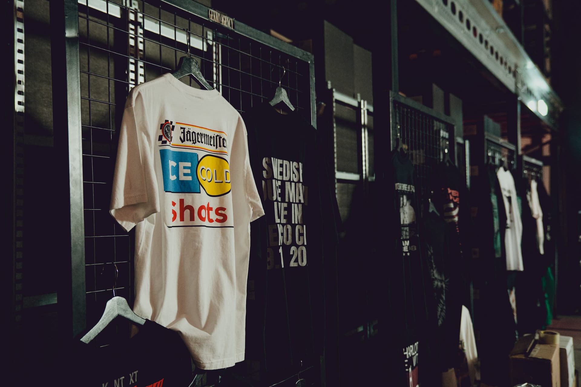 Client tees on hangers along a wire merch wall in the warehouse, with a white Jägermeister Ice Cold Shots tee in front and rows of shirts receding down the aisle.