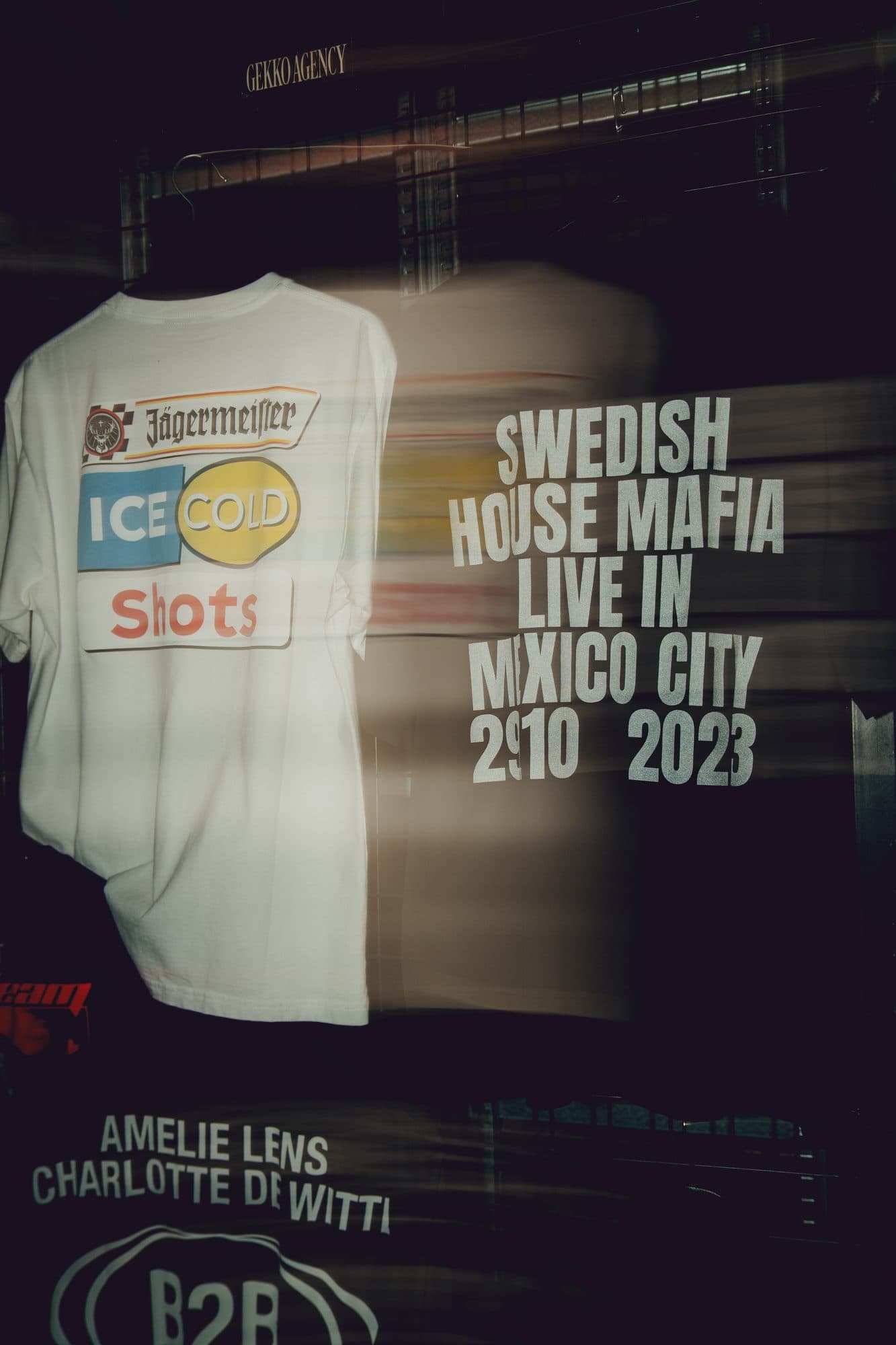 Long exposure at the merch wall: a white Jägermeister Ice Cold Shots tee on a hanger, a blurred tee printed Swedish House Mafia Live in Mexico City 2023 beside it and a black Amelie Lens and Charlotte de Witte B2B tee below, under a small GEKKO AGENCY sign.
