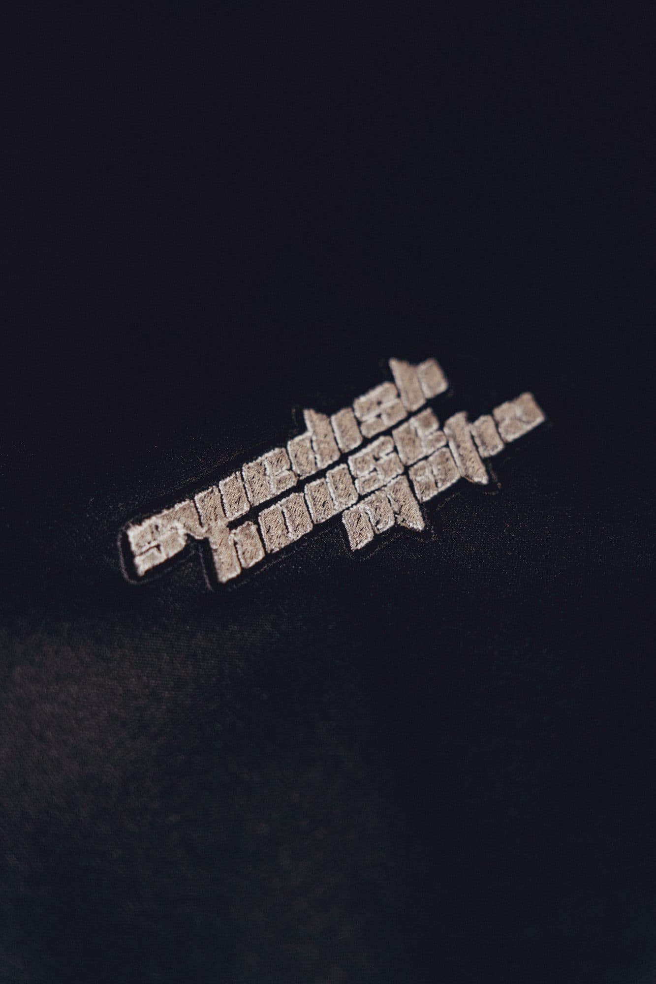 Light grey embroidered Swedish House Mafia logo on black fabric, surrounded by dark negative space.