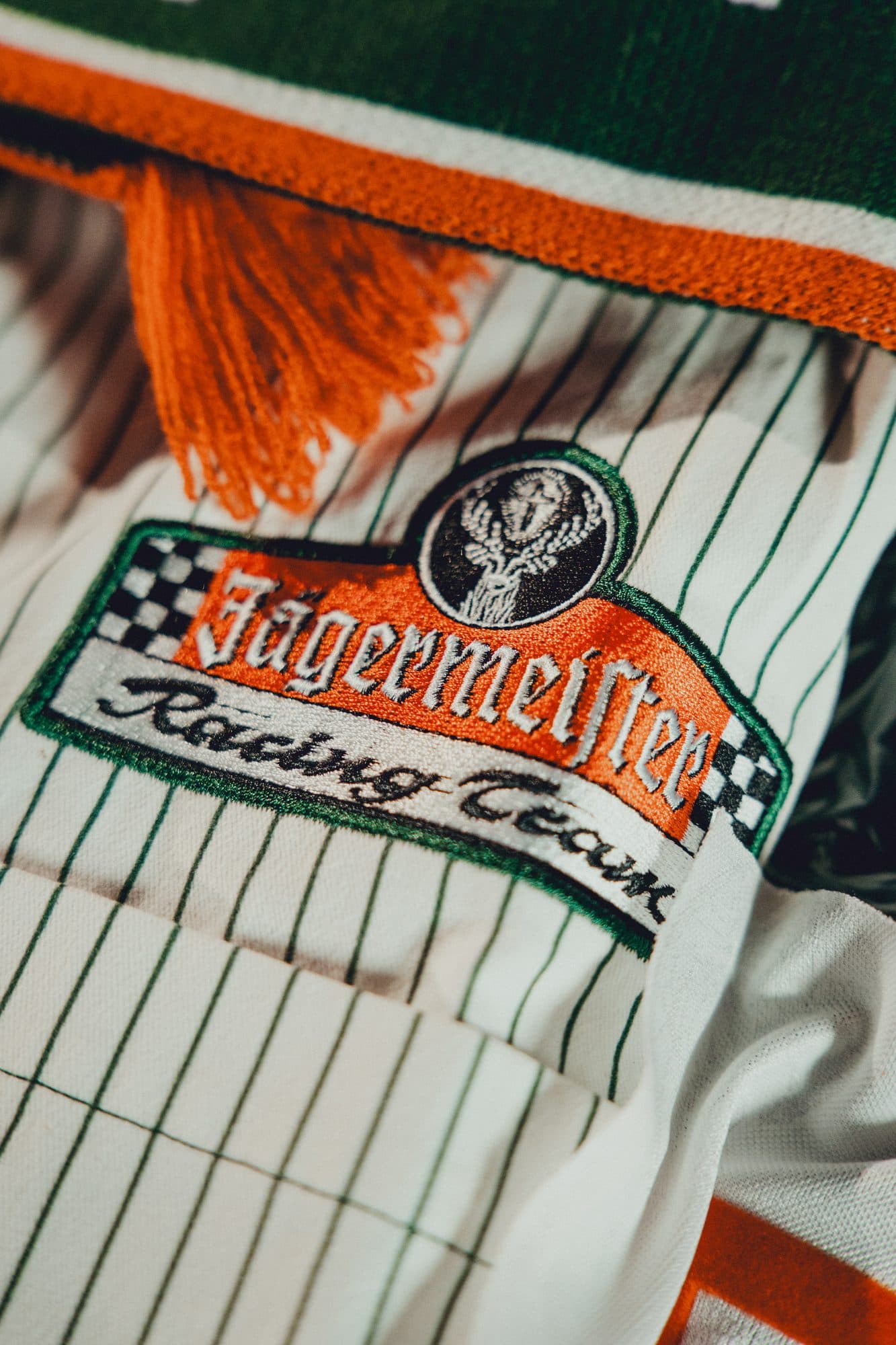 Embroidered Jägermeister Racing Team patch with a stag crest on a white shirt with green pinstripes, below the orange fringe of a scarf.