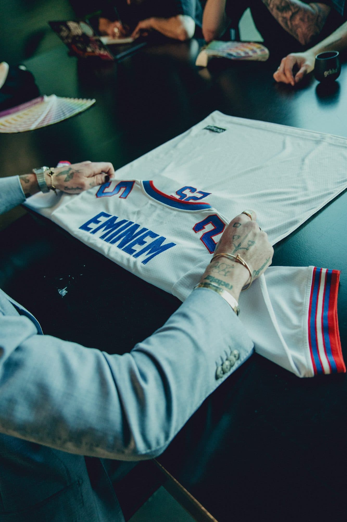 Tattooed hands straightening a white Eminem 25 jersey on the black work table, with colour swatch fans and other team members at the table behind.
