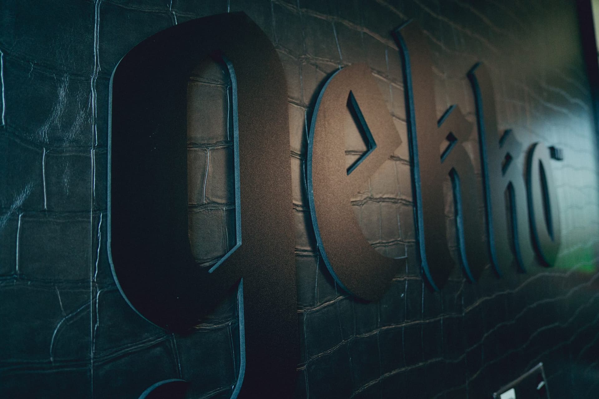 Angled view of bronze-coloured three-dimensional GEKKO letters on a black crocodile-embossed wall in teal light.