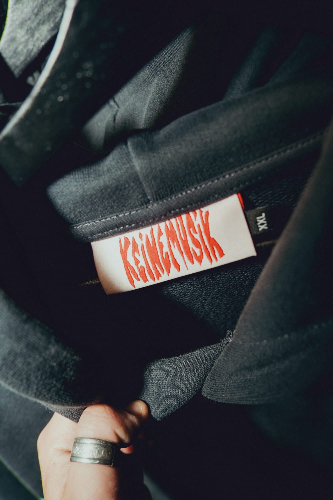 Hand with a silver ring holding open a washed black hoodie to show a red-on-white woven Keinemusik neck label and an XXL size tag.