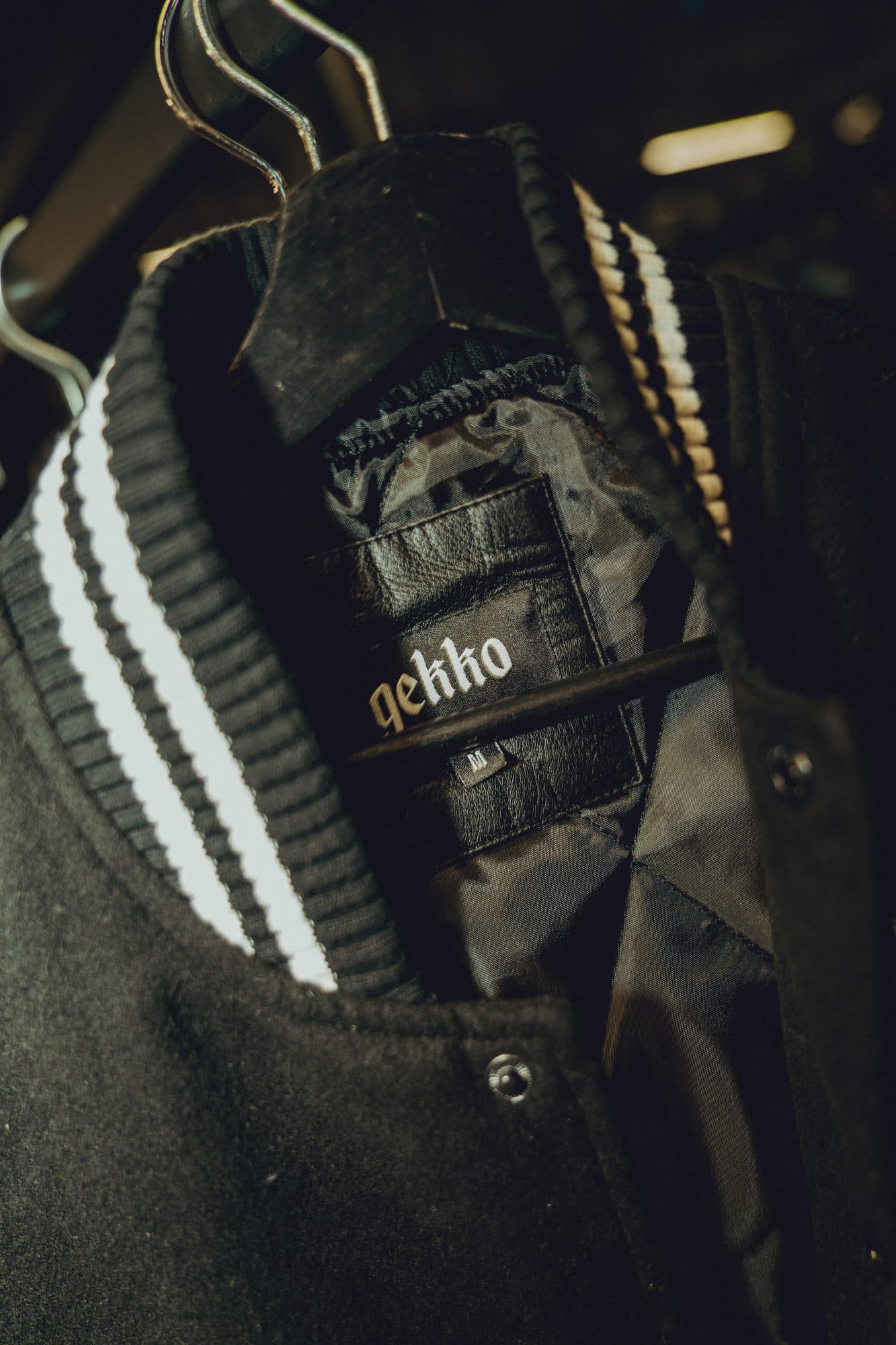 Inside of a black wool varsity jacket on a hanger, showing a black leather GEKKO label, a size tag and a striped rib collar.