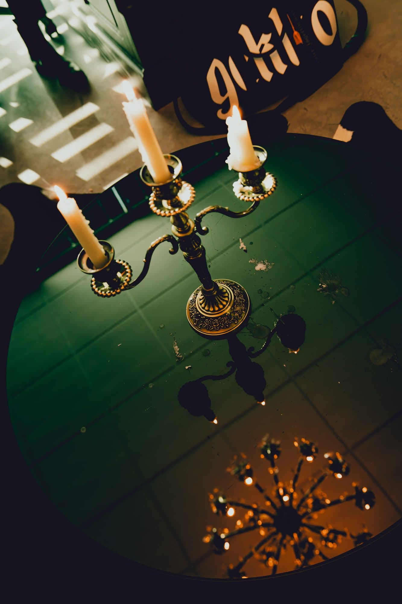 Lit brass candelabra seen from above on a round green mirrored table that reflects a crystal chandelier, with a black GEKKO duffel bag behind it.