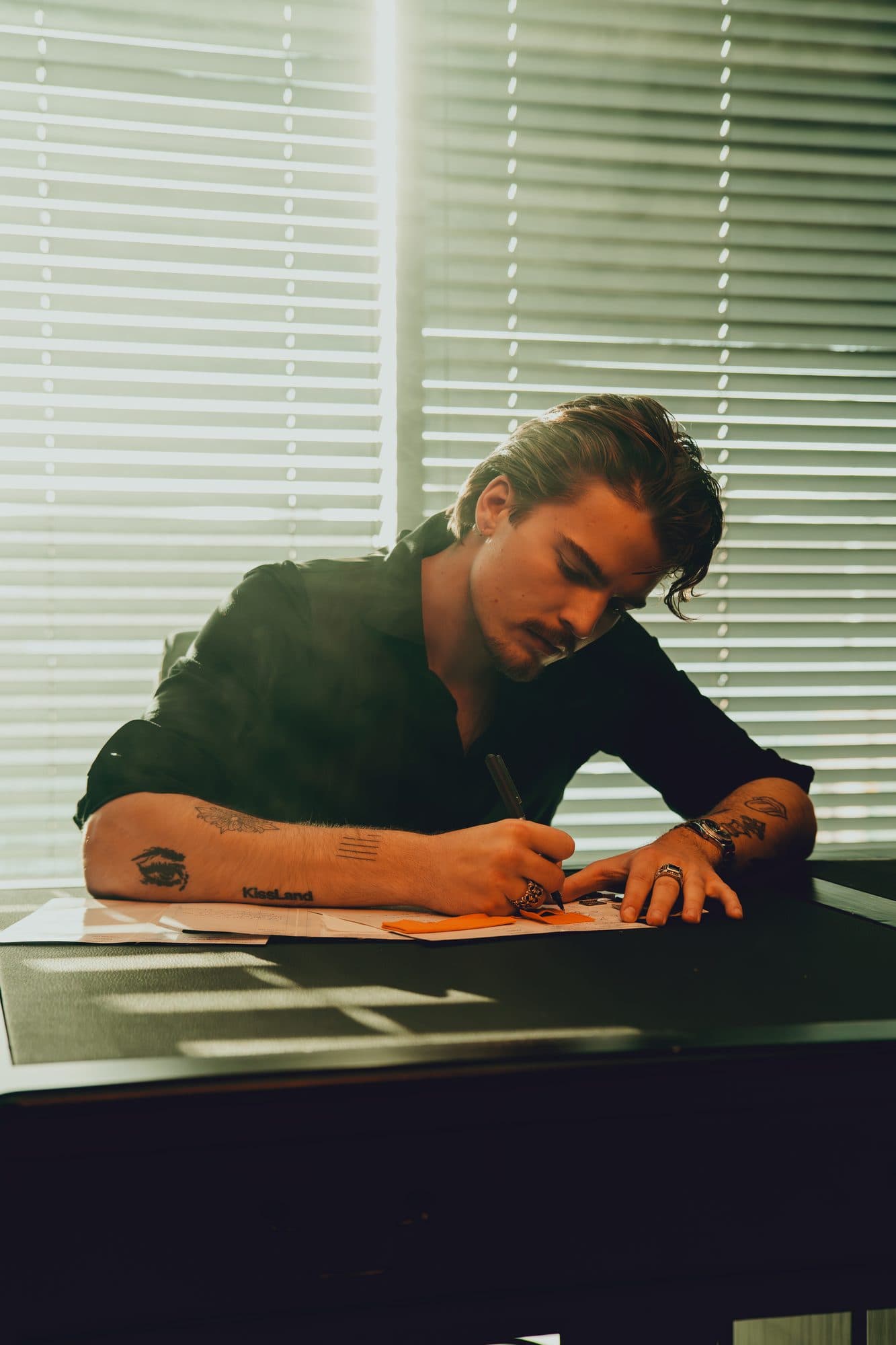 Team member sketching on tech pack sheets with orange fabric swatches at a desk, backlit by sunlight through venetian blinds.