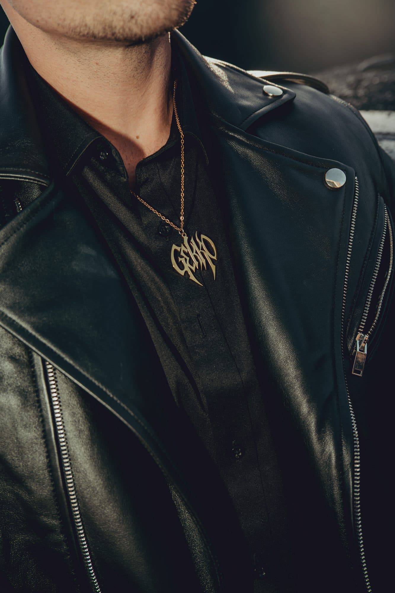 Gold GEKKO logo pendant on a chain over a black shirt and a black leather biker jacket in warm side light.