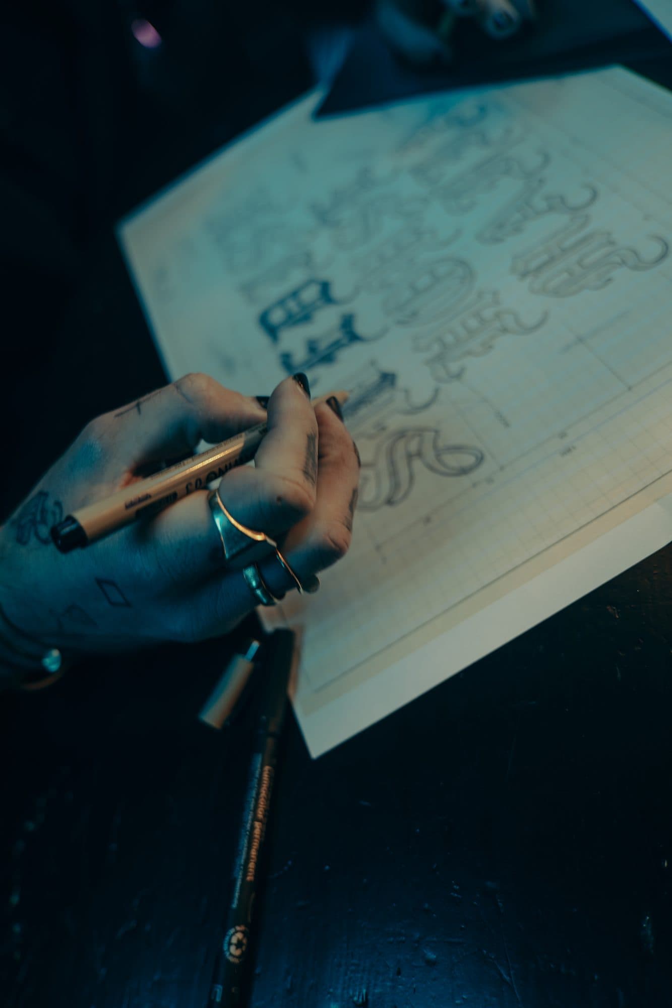 Tattooed hand with rings drawing gothic lettering with a fineliner on grid paper, a permanent marker beside the sheet.