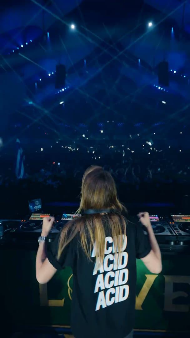 A person seen from behind at the decks with both fists raised, in a black T-shirt with ACID four times in large white letters on the back, above a green booth front with large gold letters and facing a dark arena crossed by blue laser beams; the film is one take of the dance.