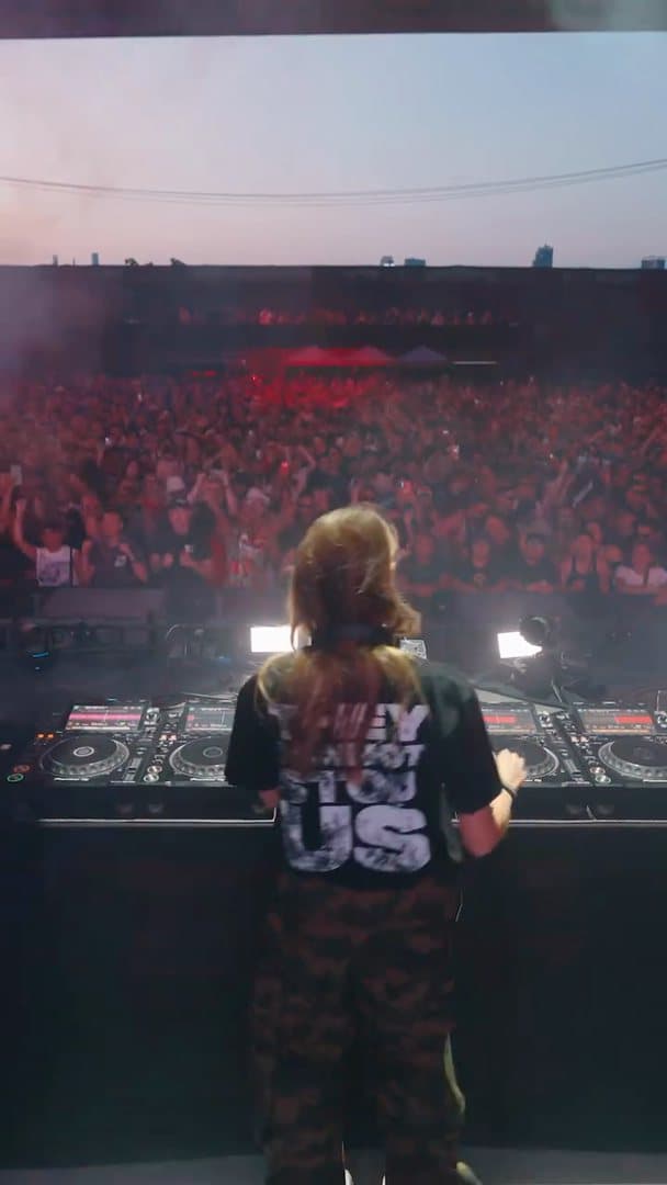A person seen from behind at the decks at dusk, in a black T-shirt with a large distressed white back print ending in US and camouflage trousers, facing a packed crowd in red light; the film also shows a stage screen with a large face and CHARLOTTE in white letters above the crowd, and the person dancing at the decks, turning sideways.