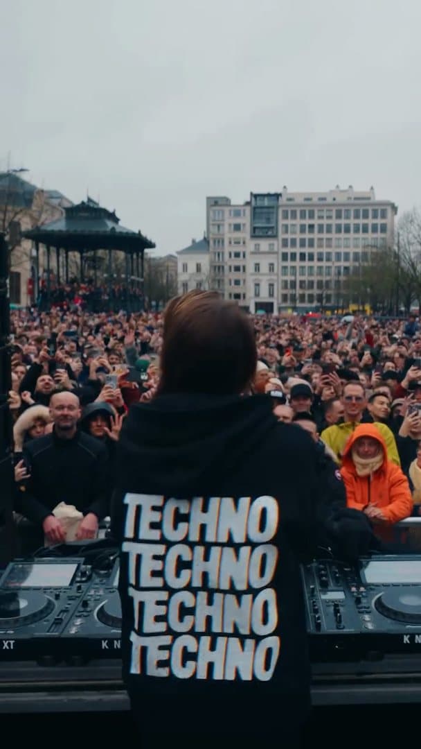 A person seen from behind at the decks in a black hoodie with TECHNO four times in large white letters on the back, facing a packed city square with a bandstand under a grey sky; the film goes on to the crowd filling the square and the person facing the camera with fists raised, a small white print on the chest of the hoodie.
