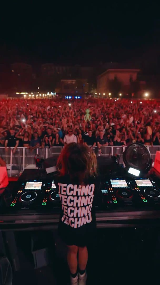 A person seen from behind at the decks in a black T-shirt with TECHNO four times in large pale letters on the back, facing a packed open-air crowd at night in red light, with buildings beyond; the film is one take of the person dancing at the decks.