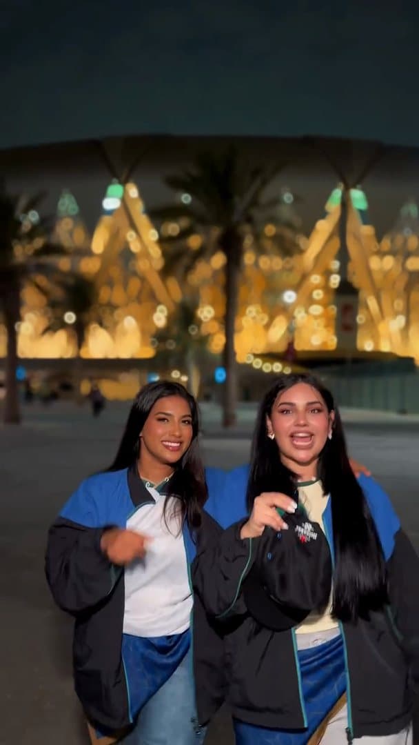 Two people in black track jackets with blue shoulders and blue patterned wraps at the hip, one holding a black cap, dancing in front of a lit stadium at night; the film is one take.