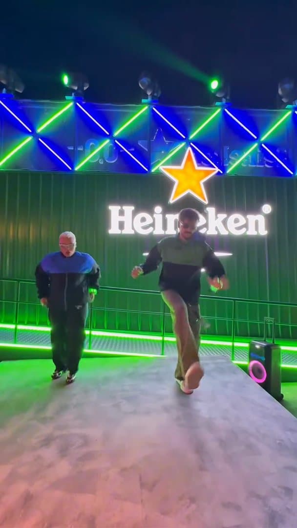 Two people in black and blue track jackets dancing on a stage in front of a lit Heineken sign with a red star, under green and blue lights; the film is one take of the dance.