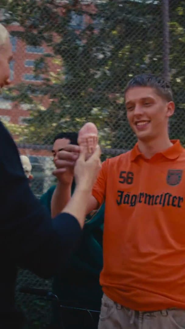 Friends on a fenced court hold up an ice cream cone together, one of them a person in the orange Jägermeister football shirt with a black 56; the film goes on to a black 56 longsleeve, orange suede sneakers, a white Jägermeister shirt with a black V-neck collar and a gold Jägermeister chain.