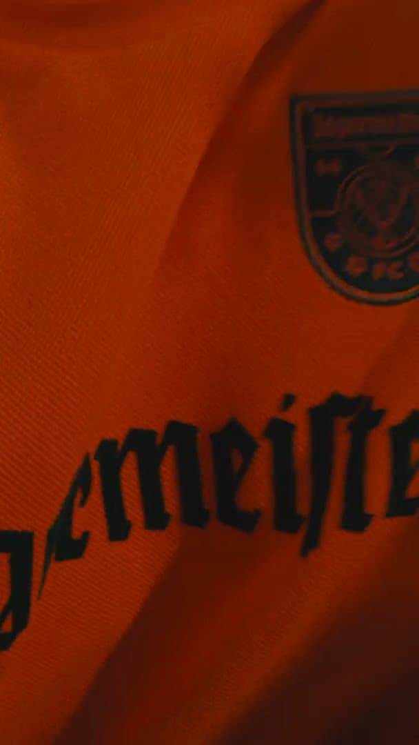 Close-up of an orange Jägermeister shirt with black gothic lettering and a crest; the film also shows a person in a black top with white Jägermeister lettering, a bottle of Jägermeister Orange and friends, one of them in the orange shirt, others in a green cap and a black hoodie, playing a stacking-block game on a rooftop.