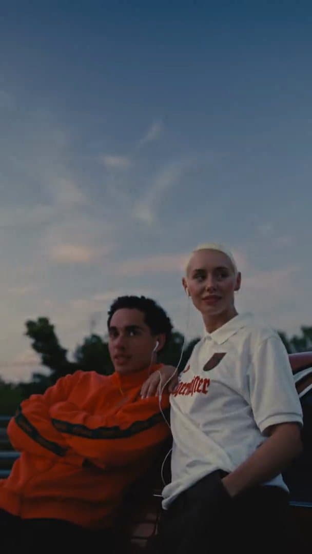 Two people, one in an orange track top with dark green stripes and one in a white Jägermeister polo shirt with red lettering, leaning on a red car under an evening sky; the film goes on to the green emblem on the orange top, a rider on a bike on a parking deck and a white 56 shirt with a basketball.