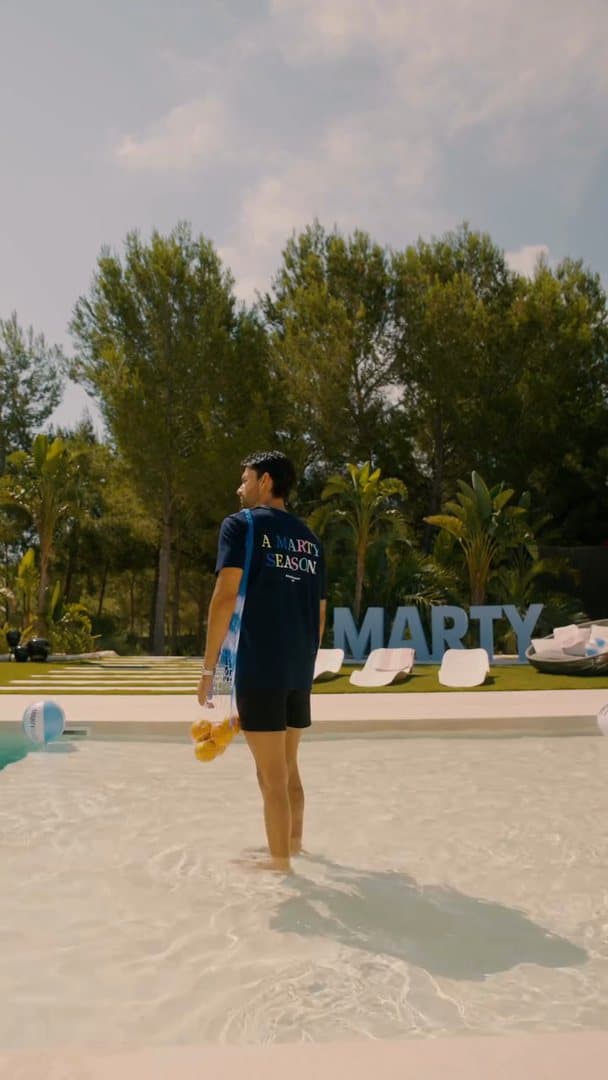 A person standing in a shallow pool in front of large light blue MARTY letters, in a navy T-shirt with A MARTY SEASON in coloured letters on the back, a net bag of yellow fruit in one hand; the film opens on a person seen from behind on a rocky shore at sunset in a navy hoodie with A MARTY SEASON on the back, looking out over a bay with boats, then shows a person in a white T-shirt with a hand fan among trees, a person in a black hoodie with a large white hexagonal emblem on a country road and a person in a white T-shirt beside a light blue open car.