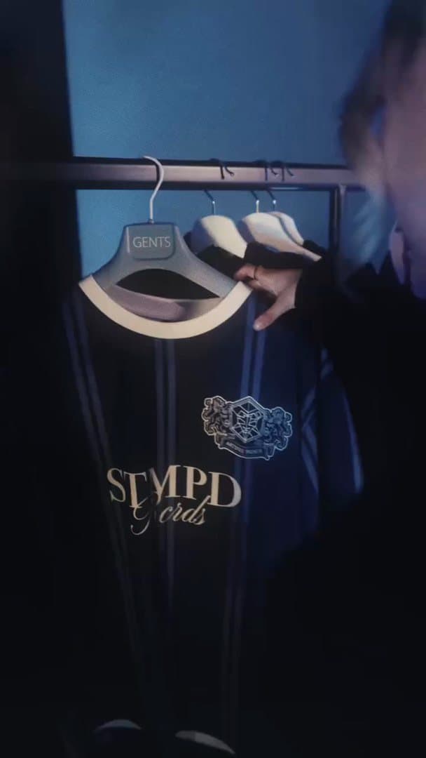 A hand taking a navy pinstripe jersey with STMPD Rcrds lettering and a crest from a clothes rail in blue light; the film goes on to a black T-shirt with a pale blue 10 and a white wireframe emblem on the back and three wireframe emblems glowing in the dark.