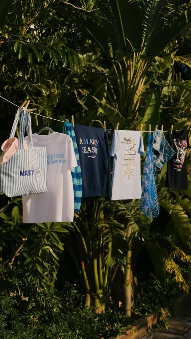 MARTY SEASON pieces pegged on a washing line in front of tropical plants: a pink hat, a blue and white striped tote bag with MARTY SEASON lettering, a white T-shirt with a light blue print, a striped towel, a navy sweatshirt with A MARTY SEASON in coloured letters, a light blue T-shirt with a signpost print, a blue mesh bag, denim shorts and a black T-shirt with a pink flower and cocktail glass print; the film moves slowly along the line as the pieces sway.