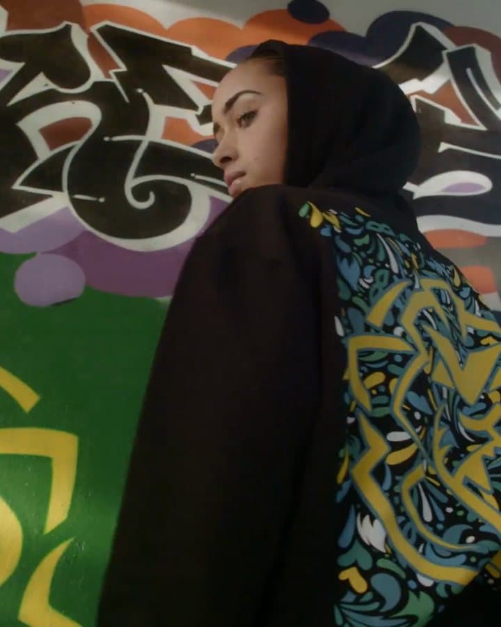 A person in a black hoodie, hood up, looking back over a shoulder in front of a graffiti wall, the back of the hoodie printed with a lion in a yellow, blue and green carnival pattern; the film also shows the person in front of a painted yellow and green lion, the hoodie from the front and a wide shot at the graffiti wall.