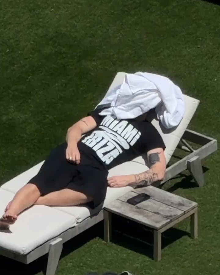 A person lying on a white sun lounger on a lawn, seen from above, with a white towel over the face and a black T-shirt with MIAMI and IBIZA in large white letters; the film zooms out over a garden of white loungers, cabanas and palm trees to a pool and the beach, while a second person walks up to the lounger.