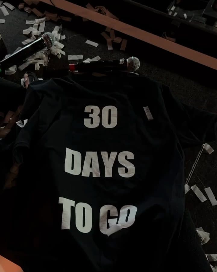 A black T-shirt printed 30 DAYS TO GO in large white letters on a stage among white confetti and microphones; the film opens on a black T-shirt printed SHM LIVE ULLEVI lying on top of it, which a pair of hands lifts away.