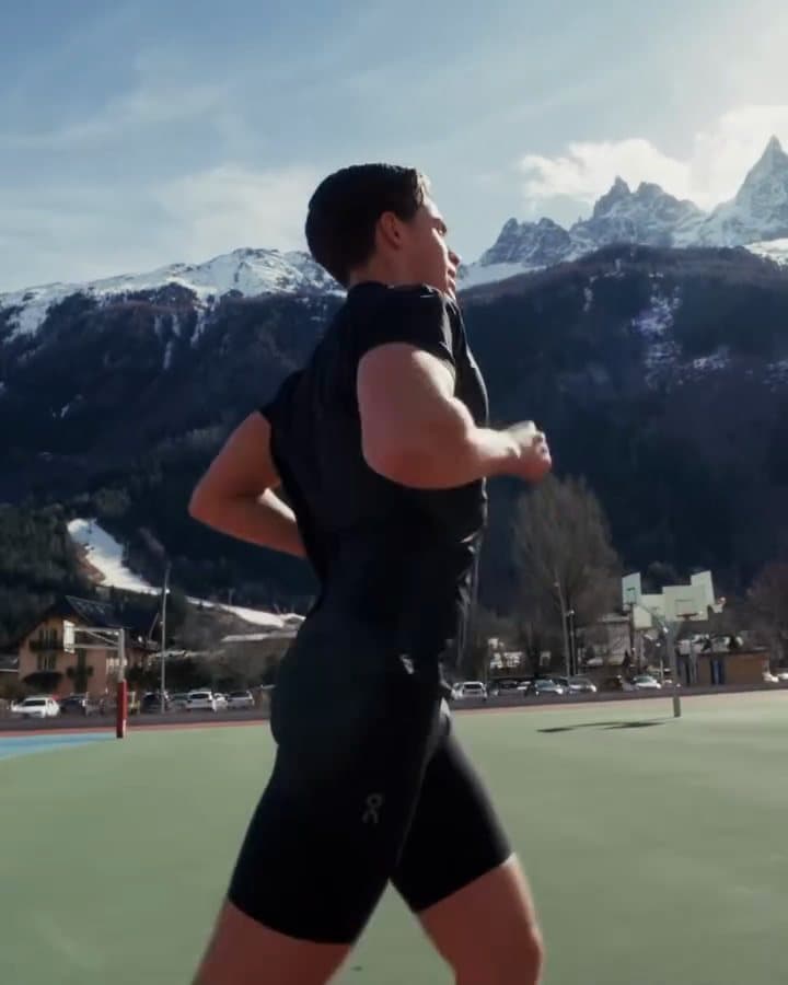 A person running in a black short-sleeved T-shirt and black shorts on a green sports ground below snowy mountains; the film also shows running shoes on a red track, the person running along a wire fence and, seen from behind, across a blue court with basketball hoops.